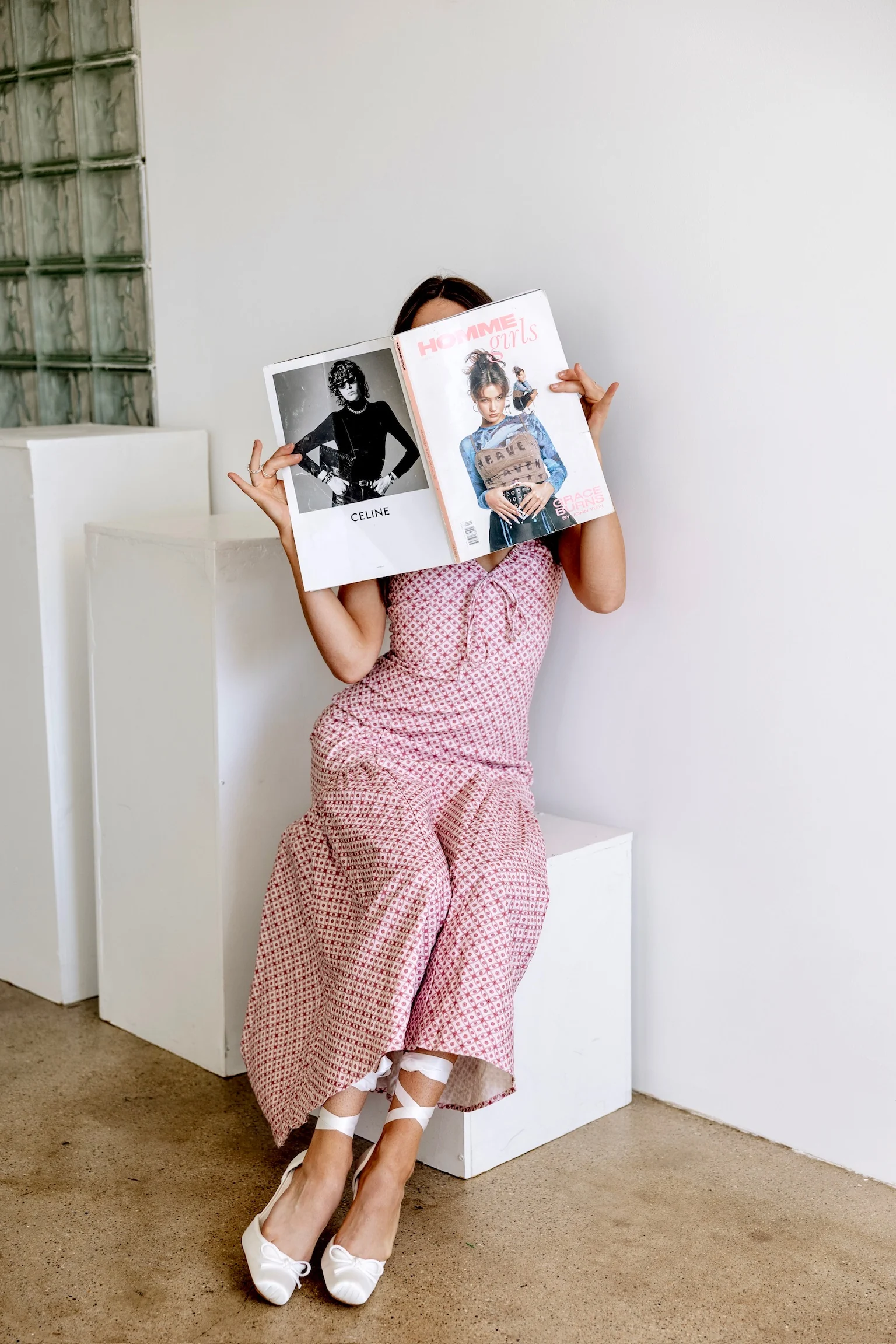 Kaylin seated on a white block in a pink dress, reading a fashion magazine.